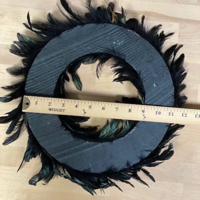 Peut inclure: Une couronne noire circulaire orn&eacute;e de plumes sombres, dot&eacute;e d'une base noire plate et textur&eacute;e. Une r&egrave;gle en bois Westcott est plac&eacute;e au centre, indiquant que la couronne a un diam&egrave;tre d'environ 30,5 cm d'un bord &agrave; l'autre.