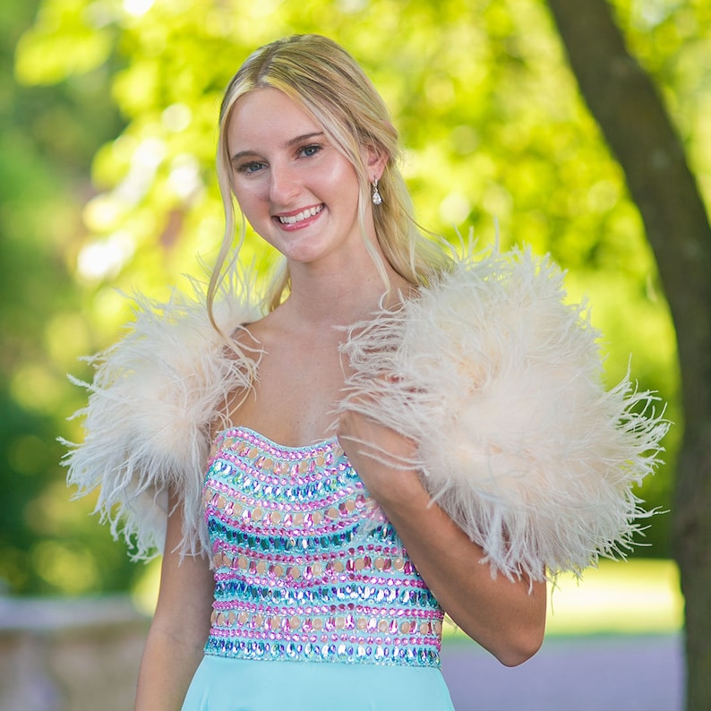 May include: A young woman wearing a light blue dress with a beaded bodice and a white feather boa. She is smiling and looking at the camera.