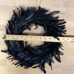 Peut inclure: Une couronne circulaire faite de plumes noires, plac&eacute;e derri&egrave;re une r&egrave;gle en bois Westcott qui montre que l'objet a un diam&egrave;tre d'environ 30 cm. Les plumes sont superpos&eacute;es et denses, cr&eacute;ant un aspect textur&eacute;, sombre et arrondi.