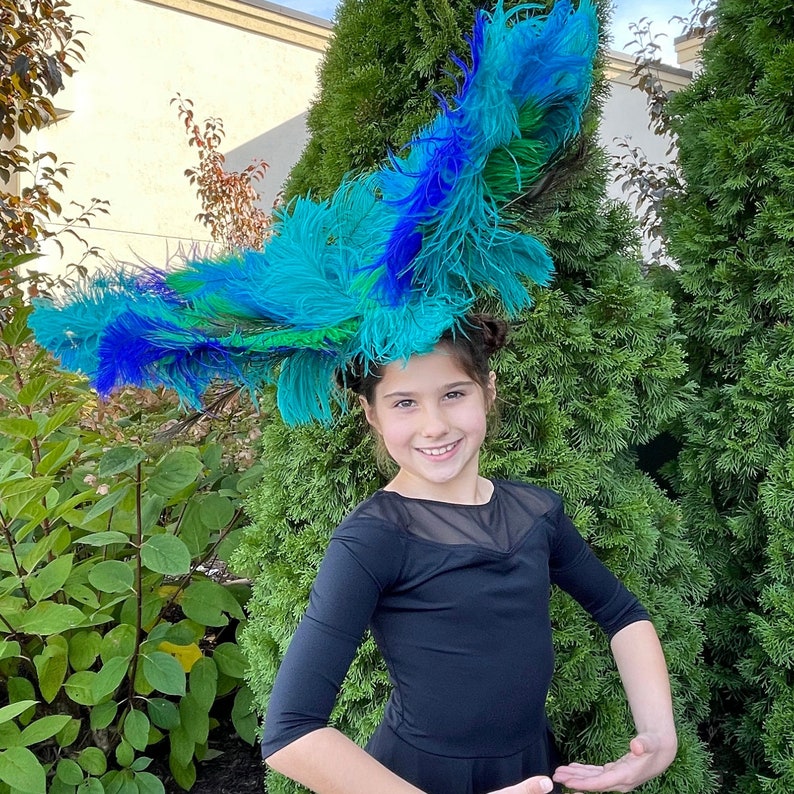 May include: A young person wearing a black dress with a large blue and green feather headdress. The headdress is reminiscent of a peacock's tail feathers.