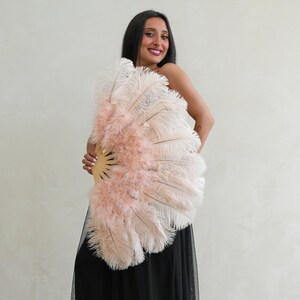 Puede incluir: Una mujer sostiene un gran abanico de plumas rosa y blanco delante de ella. El abanico est&aacute; hecho de plumas suaves y esponjosas y tiene un mango de madera.