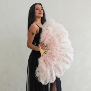 Puede incluir: Una mujer con un vestido negro sostiene un gran abanico de plumas rosa y blanco. El abanico tiene un mango de madera y est&aacute; decorado con plumas rosas.
