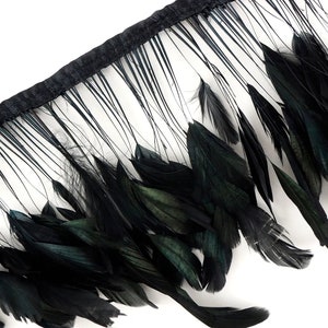 May include: Close-up of a black feather fringe trim. The trim features a black fabric base with long, thin, black feathers and larger, more substantial black feathers with hints of green. The feathers are arranged in a layered pattern.