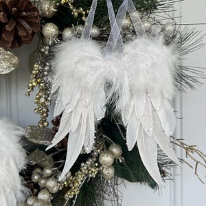 Może przedstawiać: Białe skrzydła anioła z piór na tle dekoracyjnego wieńca. Skrzydła wykonane są z miękkich, białych piór i ozdobione małymi, błyszczącymi koralikami. Wieniec zawiera szyszki, srebrne i złote ozdoby oraz zieleń.