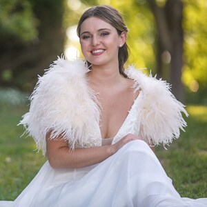 May include: A bride wearing a white wedding dress with a white feather shawl. She is sitting on a grassy lawn and smiling at the camera.