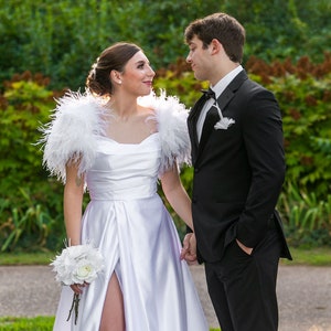 May include: A couple in formal attire, the woman wearing a white satin dress with feathered sleeves and a bouquet of white flowers, the man wearing a black tuxedo with a white bow tie.