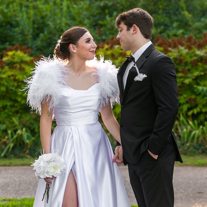 May include: A couple in formal attire, the woman wearing a white satin dress with feathered sleeves and a bouquet of white flowers, the man wearing a black tuxedo with a white bow tie.