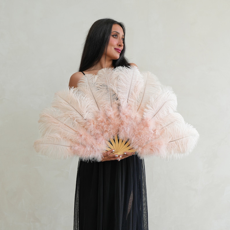 Puede incluir: Una mujer sostiene un gran abanico de plumas rosa y blanco frente a ella. El abanico est&aacute; hecho de plumas de avestruz y tiene un mango de madera.