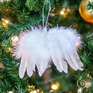 May include: White feather angel wings ornament hanging on a green Christmas tree. The wings are made of soft, fluffy feathers and have a delicate, ethereal look.