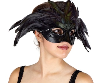 May include: A black masquerade mask with feathers. The mask has a black and green design with a bird-like beak. The mask is worn by a person with a black strap.