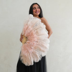 Puede incluir: Una mujer sostiene un gran abanico de plumas rosa y blanco. El abanico est&aacute; hecho de plumas suaves y esponjosas y tiene un mango de madera. La mujer lleva un vestido negro.