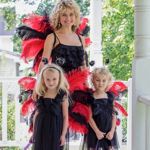 May include: Three people dressed in black and red costumes with feather wings. The woman in the middle is wearing a black dress with red and black feathers. The two girls on either side are wearing black dresses with red and black feathers. They are all smiling.