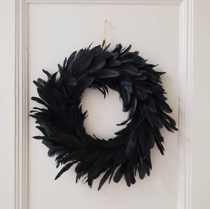 Peut inclure: Une couronne circulaire compos&eacute;e de nombreuses plumes noires superpos&eacute;es. La couronne pr&eacute;sente un aspect dense et textur&eacute; avec un centre creux et est suspendue par une fine ficelle de couleur claire fix&eacute;e au bord sup&eacute;rieur du cadre.