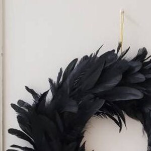Peut inclure: Une couronne circulaire compos&eacute;e de nombreuses plumes noires superpos&eacute;es. La couronne est suspendue par un fin crochet en fil m&eacute;tallique dor&eacute; vertical fix&eacute; sur le dessus, cr&eacute;ant un aspect textur&eacute; et superpos&eacute; d'une couleur sombre uniforme.