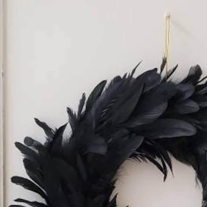 Peut inclure: Une couronne circulaire compos&eacute;e de nombreuses plumes noires superpos&eacute;es. La couronne est suspendue par un fin crochet en fil m&eacute;tallique dor&eacute; vertical fix&eacute; sur le dessus, cr&eacute;ant un aspect textur&eacute; et superpos&eacute; d'une couleur sombre uniforme.