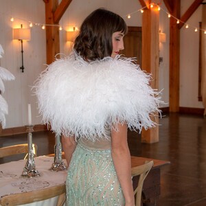 May include: A white feather boa shawl draped over a person's shoulders. The boa is made of soft, fluffy feathers and is a luxurious accessory.