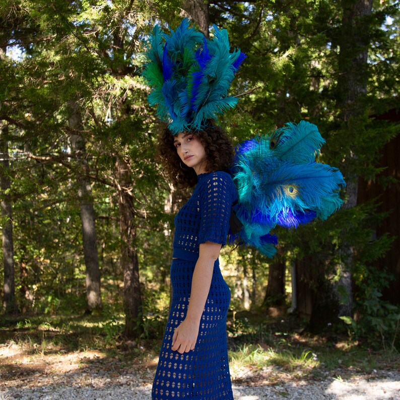 May include: A woman wearing a blue crochet dress and a blue and green feather headdress. The headdress is large and dramatic, and the woman is standing in a forest setting.