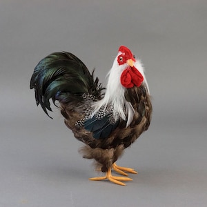 May include: A decorative rooster figurine with a white head, red comb and wattle, and a brown and white speckled body. The tail feathers are black and green. The rooster has yellow feet and is set against a gray background.