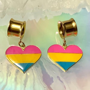 May include: A pair of gold-colored ear gauges with heart-shaped pendants. The pendants feature the pansexual pride flag colors: pink, yellow, and blue. The gauges are designed for stretched ear piercings.