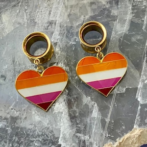 May include: A pair of gold-colored ear gauges with a heart-shaped charm. The charms are painted with a lesbian pride flag design in orange, white, pink, and red.