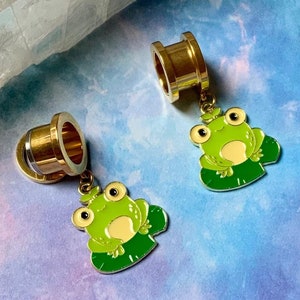 May include: A pair of gold-colored ear gauges with frog charms. The frogs are green with white eyes and are sitting on lily pads.