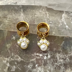 May include: A pair of gold-toned ear gauges with a dangling pearl charm. Each charm features a large pearl surrounded by smaller pearls.