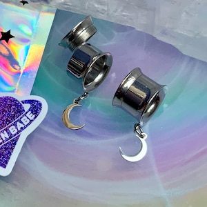 May include: Three silver-colored ear gauges with dangling gold-colored crescent moons. The gauges are displayed on a holographic surface with a sticker that says "PINK ALIEN BABE" and a cartoon alien head.