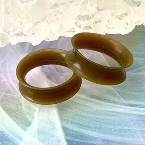 May include: Two brown silicone ear gauges, also known as flesh tunnels, on a white and blue background.