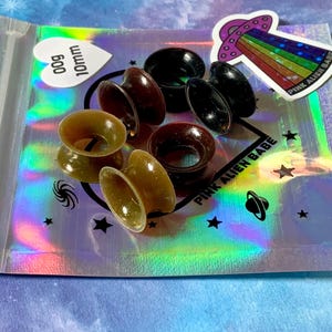 May include: A close-up of a silver holographic bag with a zipper closure. The bag contains a set of six brown and black silicone ear gauges, each measuring 10mm in diameter. The bag also features a sticker with a pink alien and the text "Pink Alien Babe".