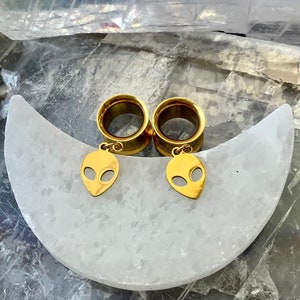 May include: A pair of gold-colored ear gauges with alien face charms hanging from them. The gauges are displayed on a white crescent moon-shaped surface.