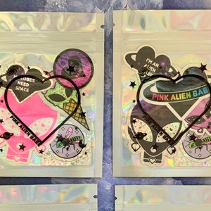 May include: Four clear plastic bags with holographic designs, each containing a set of four stickers with an alien theme. The stickers feature pink, green, black, and white colors and include phrases like "Pink Alien Bab", "My Lobes Need Space", and "I'm an Alien".