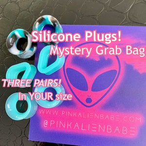 May include: A purple and pink card with an alien face graphic and the text "Silicone Plugs! Mystery Grab Bag THREE PAIRS! In YOUR size WWW.PINKALIENBABE.COM @PINKALIENBABE". The card is surrounded by turquoise silicone ear plugs.