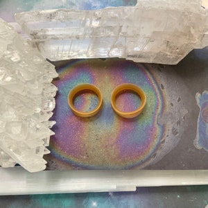 May include: Two brown, round, acrylic ear gauges on a background of iridescent, rainbow-colored oil slicks and white crystal clusters.