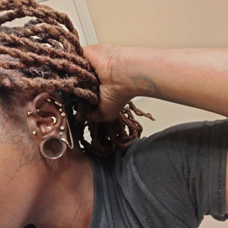 May include: A person with dreadlocks has multiple ear piercings, including a large gauge earlobe piercing with a brown plug. The earlobe piercing is adorned with a gold chain and a small silver crescent moon charm.