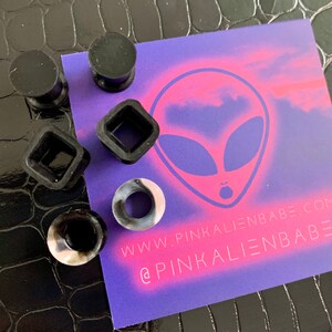 May include: A set of five silicone ear gauges in various shapes and colors, including black, white, and gray. The gauges are displayed on a purple background with an alien graphic and the text "www.pinkalienbabe.com @pinkalienbabe".