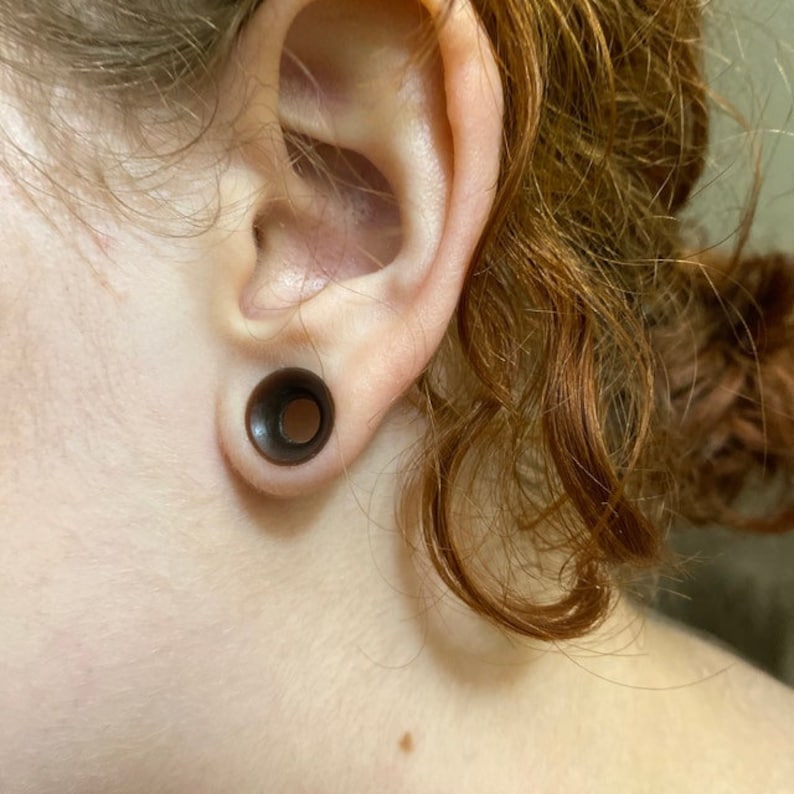 May include: A close-up of a person's ear with a black plug-style earring. The earring is a simple, round design with a smooth surface.
