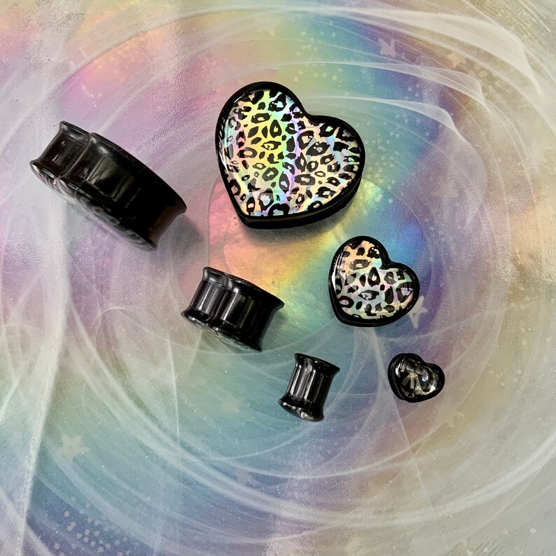 May include: A set of black acrylic ear gauges with a variety of sizes. The gauges are displayed on a white background with a swirl pattern. There are two heart-shaped gauges with a leopard print design.
