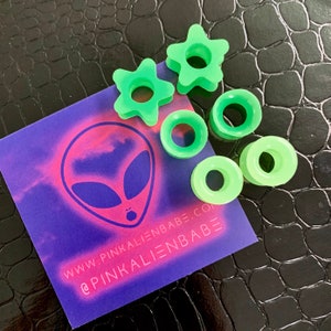May include: A set of six green glow-in-the-dark acrylic ear gauges in various shapes, including circles and stars. The gauges are displayed on a purple and pink background with an alien graphic and the text "www.pinkalienbabe.com" and "@pinkalienbabe".