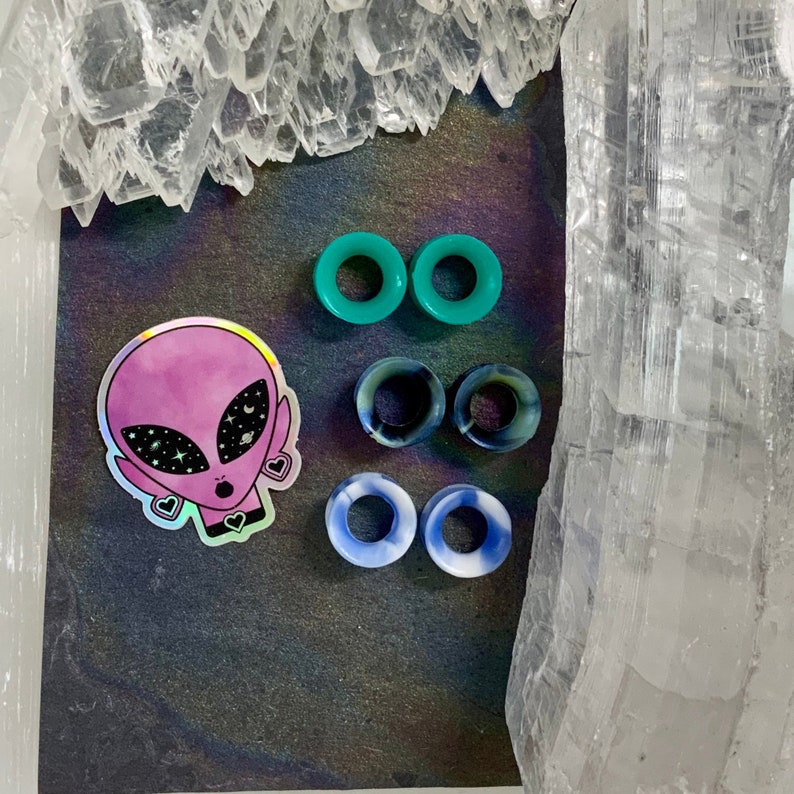 May include: A set of six silicone ear gauges in three different colors: teal, blue, and black. The gauges are arranged in pairs, with the teal gauges on top, followed by the blue gauges, and the black gauges on the bottom. The gauges are displayed on a black background with a holographic sticker of an alien with stars in its eyes.