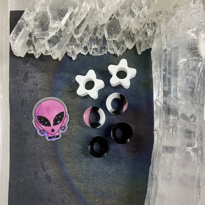 May include: A set of six ear gauges, including two white star-shaped gauges, two black and white swirl gauges, and two black gauges. The gauges are displayed on a black surface with a pink alien sticker.