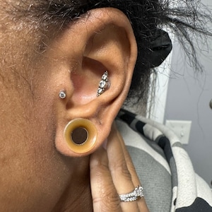 May include: A person's ear with a stretched lobe and a large, brown, silicone ear gauge. The ear also has a small, silver stud with a clear gem in the upper lobe and a silver bar with three clear gems in the middle lobe.