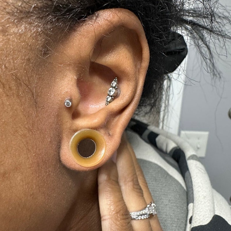 May include: A person's ear with a stretched lobe and a large, brown, silicone ear gauge. The ear also has a small, silver stud with a clear gem in the upper lobe and a silver bar with three clear gems in the middle lobe.