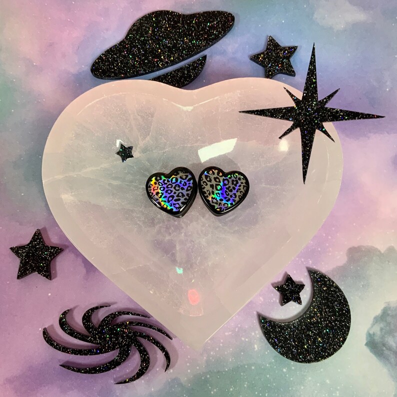 May include: Two iridescent heart-shaped plugs with a leopard print design, displayed on a white heart-shaped selenite stone. The stone is surrounded by black glitter stars and a crescent moon.