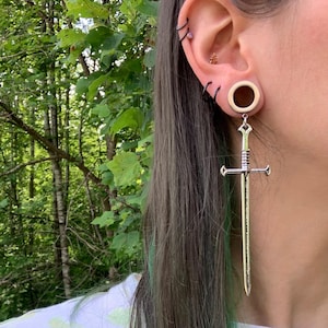 May include: A silver-toned sword-shaped earring pendant hanging from a circular ear gauge. The sword features a detailed hilt, crossguard, and a long, straight blade. Two thin black hoop earrings are also visible in the upper ear cartilage.