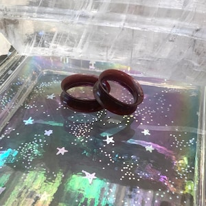 May include: Two dark purple ear gauges, also known as flesh tunnels, are displayed on a glittery surface. The gauges are made of a smooth, shiny material.