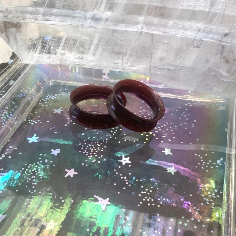 May include: Two dark purple ear gauges, also known as flesh tunnels, are displayed on a glittery surface. The gauges are made of a smooth, shiny material.