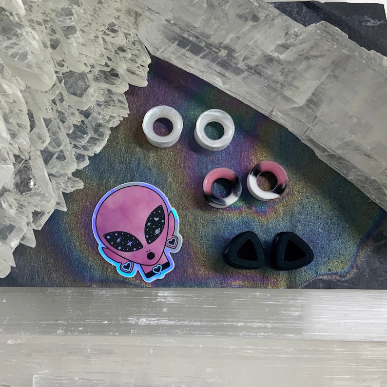 May include: A set of six ear gauges in various colors and shapes, including black, white, pink, and clear. The gauges are made of acrylic and silicone. There is also a sticker of an alien with a galaxy design.