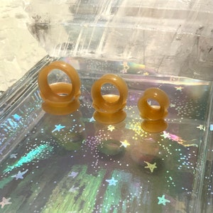 May include: Three brown rubber ear gauges of different sizes. The gauges are on a sparkly surface.