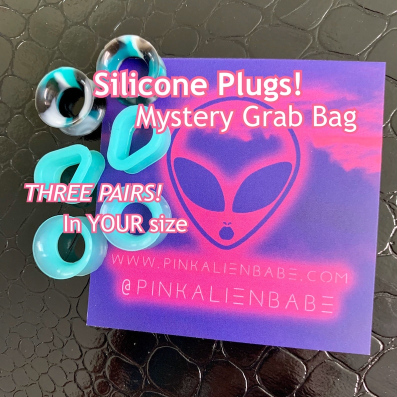 May include: A purple and pink card with an alien face graphic and the text "Silicone Plugs! Mystery Grab Bag THREE PAIRS! In YOUR size WWW.PINKALIENBABE.COM @PINKALIENBABE". The card is surrounded by turquoise silicone ear plugs.