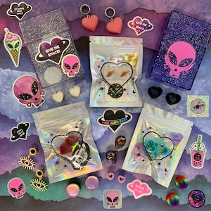 May include: A collection of colorful, glittery, and holographic packaging for jewelry and accessories. The packaging features alien and heart designs with the text "Give Me Space!" and "Pink Alien Babe".